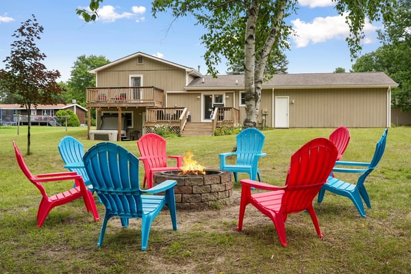Colorful Adirondack chairs arranged around a stone fire pit in the yard of a beige house with wooden decks.