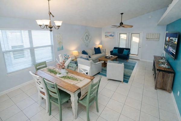 Open-plan living and dining area with tile flooring, dining table with green chairs, beige and blue seating, ceiling fan, and glass doors to balcony.